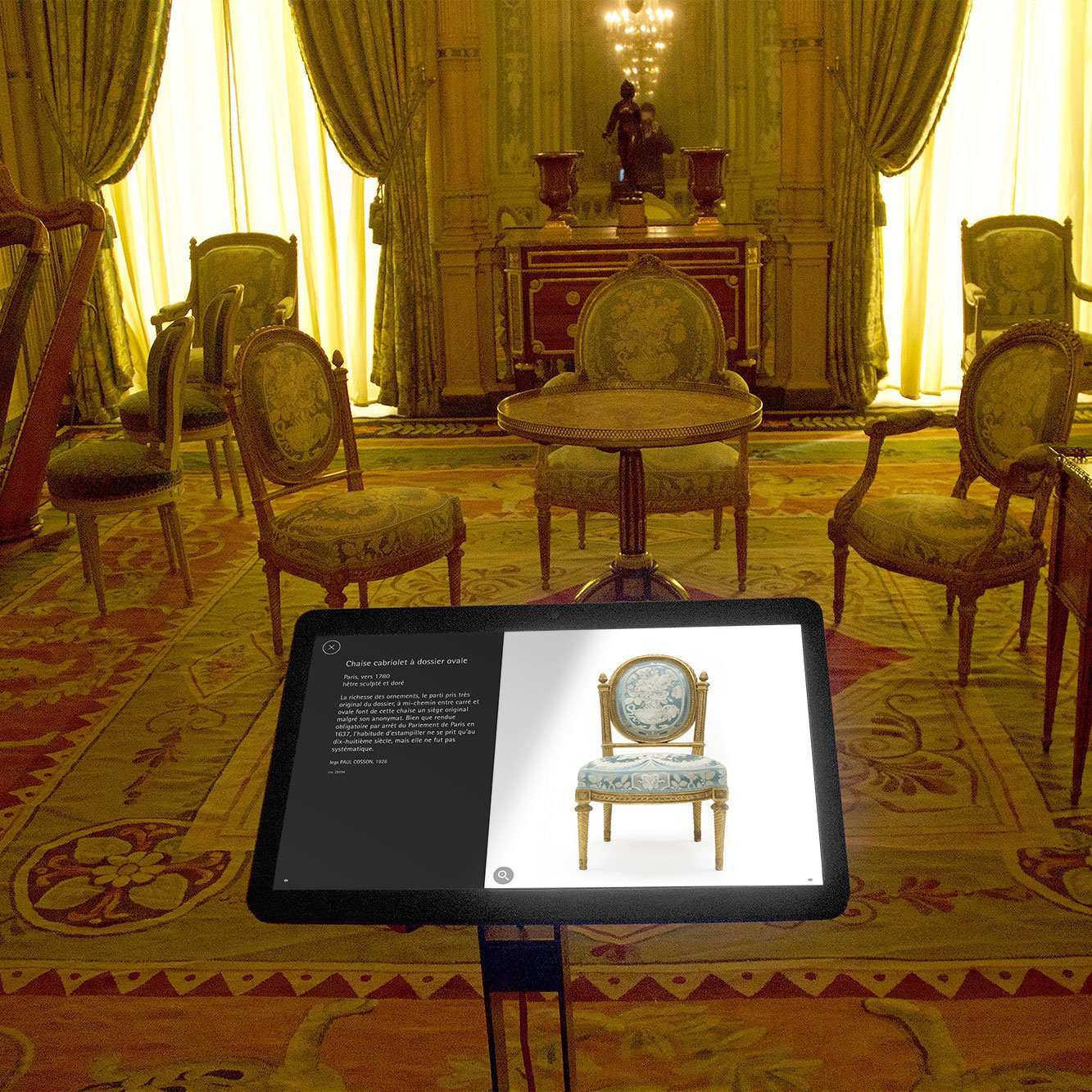 L'image montre une pièce élégante, apparemment un salon, avec plusieurs chaises ornées et une petite table en bois. Les rideaux sont tirés, laissant entrer une lumière douce qui met en valeur les détails de la décoration. Au centre, un écran interactif affiche des informations sur une chaise, ajoutant une touche moderne à cet espace traditionnel. Le sol est recouvert d'un tapis richement décoré, renforçant l'ambiance classique de la pièce.