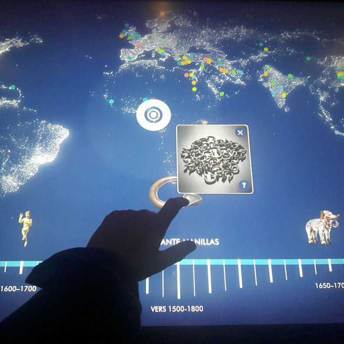L'image montre un écran interactif représentant une carte du monde, avec des points lumineux qui pourraient indiquer des emplacements géographiques ou des événements. En bas, il y a une frise chronologique qui s'étend de 1500 à 1800, avec des repères temporels. Un doigt est aperçu en train d'interagir avec l'écran, probablement pour sélectionner ou explorer des informations. Des éléments graphiques, comme une sculpture et un éléphant, sont également visibles, indiquant peut-être un lien avec des cultures ou des périodes historiques.