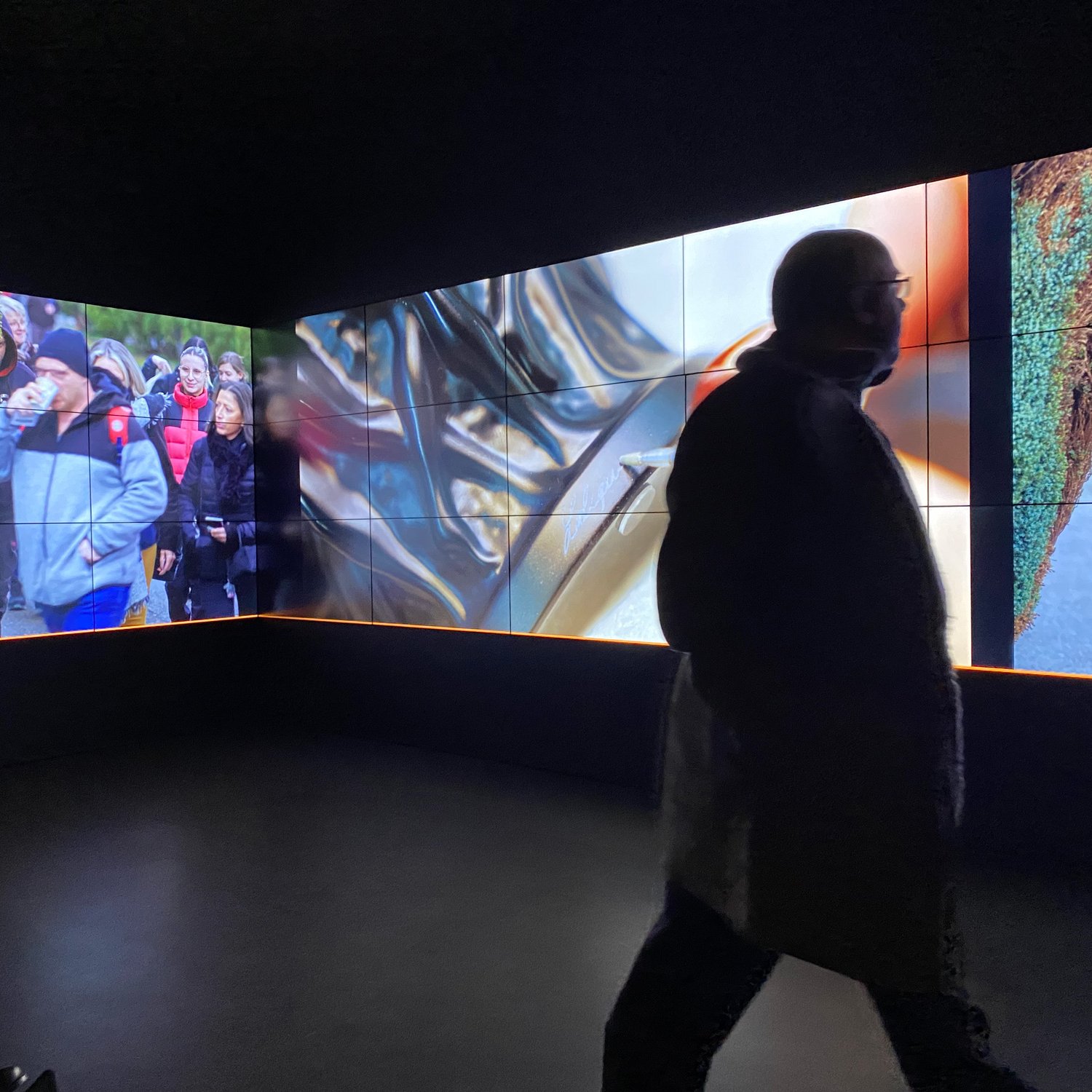 L'image montre un espace intérieur avec plusieurs écrans affichant des images variées. Sur l'un des écrans, on peut apercevoir des personnes en train de marcher, tandis que d'autres écrans présentent des motifs abstraits. Au premier plan, une silhouette d'une personne marche, créant un contraste avec les images dynamiques en arrière-plan. L'éclairage et la disposition des écrans ajoutent une atmosphère moderne et immersive à la scène.