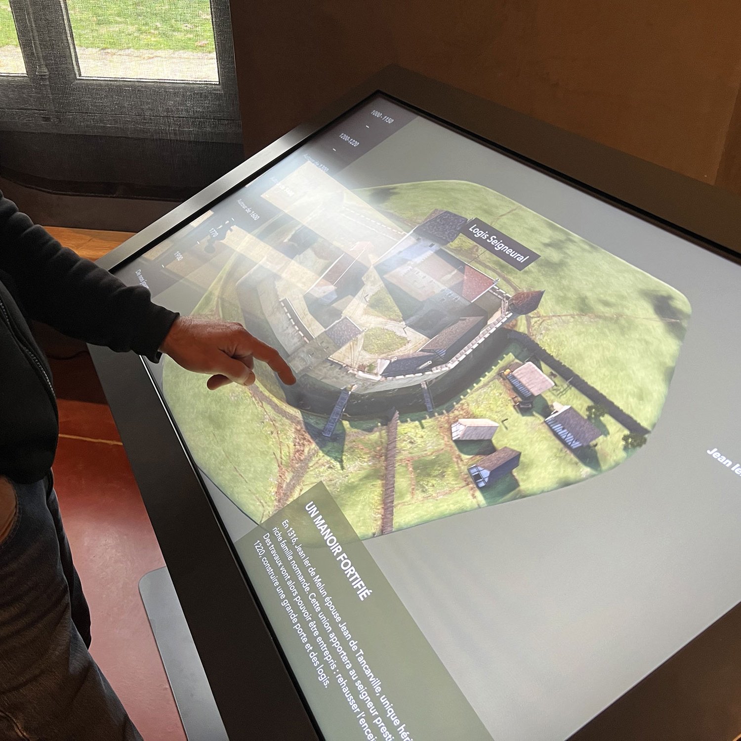 L'image montre une personne en train d'interagir avec un écran tactile. L'écran affiche une vue en 3D d'une fortification ou d'un site historique, avec des bâtiments et un espace fortifié bien définis. L'utilisateur semble examiner les détails de la présentation, en utilisant son doigt pour pointer ou interagir avec certaines parties de l'image. En arrière-plan, on peut voir des fenêtres qui laissent passer la lumière naturelle, créant une atmosphère accueillante.