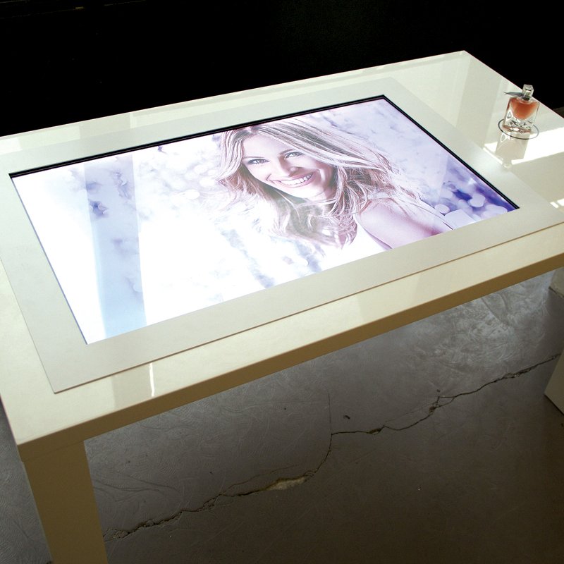 L'image montre une table moderne avec un plateau en verre. Sur ce plateau se trouve un écran tactile affichant une image, probablement d'une personne souriante. À côté de l'écran, il y a un flacon de parfum élégamment placé. La table a un design contemporain, avec des lignes épurées et une finition blanche, et elle est située dans un environnement intérieur minimaliste.