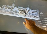 L'image montre un écran interactif représentant un bâtiment historique, probablement un hôtel de ville ou un monument, avec une vue en 3D de sa façade. On peut voir différentes parties du bâtiment mentionnées, comme "Salle du Dôme", "Salle Gothique", "Oratoire", et d'autres éléments architecturaux. Une main pointe vers le bas, probablement pour sélectionner ou interagir avec une des sections présentées sur l'écran. L'ensemble donne une impression d'exploration interactive du bâtiment.