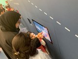 L'image montre une femme portant un hijab qui regarde un écran interactif avec une jeune fille à ses côtés. La petite fille semble interagir avec l'écran, en pointant du doigt quelque chose qui l'intéresse. En arrière-plan, d'autres enfants peuvent être aperçus, suggérant qu'ils se trouvent probablement dans un environnement éducatif ou ludique. Les murs sont peints en bleu et il y a des éléments graphiques qui évoquent un cadre amical et engageant.