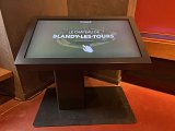 L'image montre un écran interactif installé sur un support. L'écran affiche le texte "Le Château de Blandy-les-Tours" avec une main illustrée pointant vers le texte. L'environnement semble être à l'intérieur d'un bâtiment, avec des murs en ton neutre et un sol de couleur rougeâtre.
