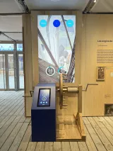 L'image montre une installation interactive dans un musée ou une exposition. Au premier plan, il y a un dispositif en bois avec une manivelle et un écran tactile. L'écran, situé au-dessus, affiche des éléments graphiques et des données, notamment des chiffres comme "100 kg" et "34" ainsi que des indications sur les valeurs. Le fond de l'installation présente un espace ouvert, peut-être avec des structures architecturales en arrière-plan. L'ensemble semble être conçu pour éduquer les visiteurs sur des moteurs ou engins mécaniques.