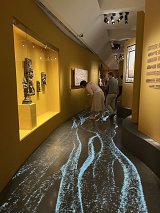 L'image montre une exposition dans un musée avec des murs peints en jaune. Au premier plan, on voit un sol illustré avec un motif lumineux qui rappelle des eaux qui s'écoulent. Deux visiteurs explorent les œuvres exposées : l'un se penche pour examiner un objet dans une vitrine, tandis que l'autre observe une œuvre accrochée au mur. L'éclairage met en valeur les sculptures et les œuvres présentes dans l'espace.