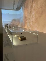 L'image montre un espace d'exposition dans un musée, avec des vitrines en verre transparent. À l'intérieur des vitrines, on peut voir quelques objets exposés, dont une boîte en bois et d'autres éléments, éclairés par un éclairage doux. En arrière-plan, un mur est orné d'une projection de paysage, probablement une représentation de la nature ou d'une scène historique, ajoutant une ambiance visuelle à l'exposition. Les textes explicatifs sont placés sur la surface en face des vitrines.