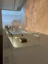 L'image montre un espace d'exposition dans un musée, avec des vitrines en verre transparent. À l'intérieur des vitrines, on peut voir quelques objets exposés, dont une boîte en bois et d'autres éléments, éclairés par un éclairage doux. En arrière-plan, un mur est orné d'une projection de paysage, probablement une représentation de la nature ou d'une scène historique, ajoutant une ambiance visuelle à l'exposition. Les textes explicatifs sont placés sur la surface en face des vitrines.