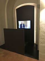 L'image montre un espace minimaliste avec un mur noir, sur lequel est fixé un écran de télévision. L'écran diffuse une vidéo d'un homme qui parle, avec des sous-titres en français. Il y a une petite plateforme ou un siège devant l'écran, et l'ensemble de l'installation est situé dans un environnement aux murs blancs et au sol en carrelage. L'atmosphère semble moderne et sobre.