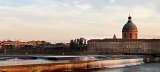 L'image montre un paysage urbain au crépuscule, avec une rivière et des chutes d'eau en premier plan. À l'arrière-plan, on peut apercevoir un bâtiment avec une grande coupole, flanqué d'autres bâtiments de style architectural classique. Un pont est également visible, traversant la rivière. Les couleurs du ciel évoquent une atmosphère tranquille, créant un contraste agréable avec l'architecture environnante.