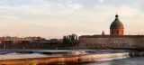 L'image montre un paysage urbain au crépuscule, avec une rivière et des chutes d'eau en premier plan. À l'arrière-plan, on peut apercevoir un bâtiment avec une grande coupole, flanqué d'autres bâtiments de style architectural classique. Un pont est également visible, traversant la rivière. Les couleurs du ciel évoquent une atmosphère tranquille, créant un contraste agréable avec l'architecture environnante.
