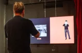 Sur l'image, on voit un homme de dos, qui semble être en train de regarder un écran. L'écran est divisé en deux parties : à gauche, une vidéo en noir et blanc capture une scène d'un événement, et à droite, une animation représentant un personnage en costume. Il y a également un chronomètre qui indique du temps. L'ambiance est moderne, probablement dans un cadre de présentation ou de performance.