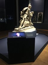 L'image présente une sculpture en marbre, représentant une figure équestre, probablement un personnage historique ou mythologique. La sculpture est placée sur une base, et à proximité, il y a un écran interactif qui fournit des informations supplémentaires. L'environnement semble être une exposition muséale avec des murs sombres, mettant en valeur l'œuvre. On peut également apercevoir d'autres sculptures en arrière-plan, suggérant un espace dédié à l'art.