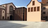 L'image montre un espace architectural avec plusieurs bâtiments. À gauche, il y a une structure qui semble plus ancienne, avec des arches et des fenêtres en hauteur, probablement en pierre. À droite, un bâtiment moderne présente une façade lisse de couleur claire, avec des volets en bois. Entre les deux bâtiments, une cour pavée est visible, sous un ciel partiellement nuageux. L'ensemble crée un contraste intéressant entre l'architecture traditionnelle et contemporaine.