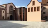 L'image montre un espace architectural avec plusieurs bâtiments. À gauche, il y a une structure qui semble plus ancienne, avec des arches et des fenêtres en hauteur, probablement en pierre. À droite, un bâtiment moderne présente une façade lisse de couleur claire, avec des volets en bois. Entre les deux bâtiments, une cour pavée est visible, sous un ciel partiellement nuageux. L'ensemble crée un contraste intéressant entre l'architecture traditionnelle et contemporaine.