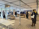 L'image montre un espace intérieur moderne, probablement une boutique ou un lieu d'exposition. Le sol est en bois, et les murs sont principalement blancs, créant une ambiance lumineuse et ouverte. Plusieurs tables sont disposées, avec des livres et des objets exposés. On peut voir des personnes qui se déplacent et discutent dans l'espace. À gauche, une personne en tenue de travail semble accueillir des visiteurs, tandis qu'au fond, d'autres personnes explorent les produits. L'éclairage est doux et met en valeur les articles exposés. L'ensemble donne une impression de convivialité et d'accessibilité.