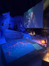 L'image montre une exposition immersive. On y voit une grande carte projetée au sol, illustrant un réseau de routes ou de parcours, probablement en lien avec un thème narratif. En arrière-plan, un écran est accroché, diffusant une scène avec des personnages, ajoutant une dimension cinématographique à l'installation. Une sculpture en forme de loup se trouve également dans l'espace, créant une atmosphère intrigante. Les visiteurs semblent interagir avec l'environnement, contribuant à l'ambiance immersive de l'exposition.