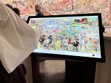 L'image montre une exposition artistique, probablement une grande fresque ou une illustration qui est affichée sur un écran numérique. On peut apercevoir des personnes autour de l'écran, avec une main pointant vers une partie de l'œuvre. La fresque semble riche en détails et en couleurs, comportant diverses scènes qui pourraient représenter des thèmes historiques ou narratifs. En arrière-plan, il y a également des murs décorés avec des œuvres d'art. L'atmosphère semble être celle d'une visite culturelle ou d'une découverte artistique.
