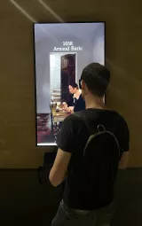 L'image montre un homme debout devant un écran numérique. L'écran affiche une œuvre d'art datant de 1658, avec le nom "Arnaud Baric" en haut. On peut voir un intérieur où deux personnes semblent assises à une table, avec un décor sobre et une lumière douce. L'homme, qui porte un sac à dos, semble observer attentivement l'image.