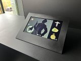 L'image montre un écran interactif monté sur une surface noire. L'écran présente une silhouette d'un homme, habillé d'un costume élégant, tenant un objet dans une main. À côté de la silhouette, plusieurs illustrations d'objets sont visibles, dont une montre de poche et d'autres éléments qui pourraient être des artefacts ou des bijoux. L'environnement semble moderne, avec des fenêtres laissant entrer la lumière naturelle.