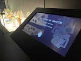 L'image montre une exposition avec des flacons de parfum sur une étagère éclairée, positionnés de manière esthétique. En avant-plan, un écran interactif affiche le texte "FLACONS DE PARFUM" et des illustrations de différents flacons. Cet écran invite à découvrir le thème des flacons de parfum à travers des informations visuelles et interactives.