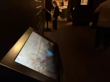 L'image montre un écran interactif placé dans une exposition ou un musée. L'écran affiche un titre qui semble être lié à une œuvre ou une thématique culturelle, bien que le texte exact ne soit pas visible. À l'arrière-plan, plusieurs personnes semblent explorer l'espace, et l'éclairage est tamisé, créant une ambiance propice à la découverte. Des panneaux d'information et des œuvres peuvent également être aperçus autour de l'écran.