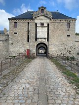 L'image montre une ancienne fortification en pierre avec une structure imposante. On peut voir un grand passage centrale encadré par des murs épais. Les fenêtres sont barrées de grilles rouges, et le toit est de style traditionnel avec des pignons. Le chemin en pavés qui mène à l'entrée est bordé de grilles, et on aperçoit une végétation discrète sur les côtés. Le ciel est légèrement nuageux, ajoutant une atmosphère historique au lieu.