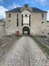 L'image montre une ancienne fortification en pierre avec une structure imposante. On peut voir un grand passage centrale encadré par des murs épais. Les fenêtres sont barrées de grilles rouges, et le toit est de style traditionnel avec des pignons. Le chemin en pavés qui mène à l'entrée est bordé de grilles, et on aperçoit une végétation discrète sur les côtés. Le ciel est légèrement nuageux, ajoutant une atmosphère historique au lieu.