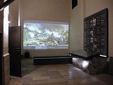 L'image montre un intérieur d'une salle, probablement un musée ou un espace d'exposition. Sur le mur, il y a une projection d'une œuvre d'art qui représente une scène de destruction avec des débris et des bâtiments en ruines, évoquant un moment dramatique. À côté de la projection, il y a un panneau d'affichage avec des informations historiques, probablement des dates et des événements. Le sol est en pierre, et il y a des éléments d'éclairage qui mettent en valeur la présentation. L'atmosphère semble à la fois éducative et réfléchie.