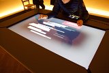 L'image montre une personne en train d'interagir avec une grande table tactile. L'écran affiche des graphiques ou des données structurées, avec des barres verticales ou des lignes. La personne utilise un doigt pour manipuler l'interface, tandis qu'une tablette est posée à côté. L'ambiance générale semble moderne et technologique, avec un éclairage doux en arrière-plan.