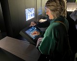 L'image montre une femme, penchée sur une tablette interactive, touchant l'écran. Elle porte un pull vert et des lunettes. En arrière-plan, on peut voir une autre personne, mais son détail n'est pas clairement visible. L'environnement semble être une exposition, avec des affichages numériques ou des projections sur les murs.