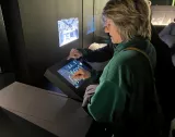 L'image montre une femme, penchée sur une tablette interactive, touchant l'écran. Elle porte un pull vert et des lunettes. En arrière-plan, on peut voir une autre personne, mais son détail n'est pas clairement visible. L'environnement semble être une exposition, avec des affichages numériques ou des projections sur les murs.