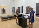 L'image montre une scène à l'intérieur d'une galerie d'art. On peut voir deux personnes, un homme et une femme, portant des masques. L'homme, habillé d'une chemise à motifs colorés et d'un short, se tient devant un écran interactif, tandis que la femme, en robe bleue, semble consulter des informations sur une autre interface. En arrière-plan, plusieurs œuvres d'art sont accrochées aux murs, se distinguant par des couleurs vives et des formes variées. La pièce est spacieuse et lumineuse, avec un sol en bois clair et un banc noir en cuir à proximité.