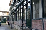 L'image montre un bâtiment dont la façade est en verre, avec des structures en métal de construction ou de rénovation visibles. Les murs sont en briques, et il y a des grandes fenêtres qui laissent entrer la lumière. Une rampe d'escalier mène à une entrée, et on peut apercevoir une cour intérieure. Les lieux semblent en pleine transformation, et quelques éléments de chantier comme des échafaudages sont présents.