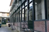 L'image montre un bâtiment dont la façade est en verre, avec des structures en métal de construction ou de rénovation visibles. Les murs sont en briques, et il y a des grandes fenêtres qui laissent entrer la lumière. Une rampe d'escalier mène à une entrée, et on peut apercevoir une cour intérieure. Les lieux semblent en pleine transformation, et quelques éléments de chantier comme des échafaudages sont présents.