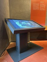 L'image montre un écran tactile moderne monté sur un socle dans un environnement intérieur. L'écran est légèrement incliné, ce qui le rend accessible pour l'interaction. À l'arrière-plan, on aperçoit un mur avec des panneaux, vraisemblablement informatifs, d'une couleur rouge. Le sol est en matériaux durs et présente une teinte rougeâtre. L'ensemble de la scène donne une impression de technologie et d'interactivité, probablement dans un musée ou un centre d'exposition.