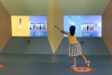 L'image montre une petite fille portant un t-shirt jaune et une jupe rayée, interagissant avec des écrans situés dans un espace coloré. Les écrans affichent des animations et des images, probablement liées à des activités ludiques ou éducatives. La fille semble engagée et concentrée sur l'activité, tandis que d'autres personnes sont visibles en arrière-plan. L'environnement est lumineux et attractif, invitant à l'exploration.