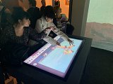 L'image montre un groupe de personnes dans un espace d'exposition, probablement dans un musée ou une galerie d'art. Deux femmes se tiennent devant un écran interactif sur une table, en train de toucher et de discuter de ce qui s'affiche. L'écran semble montrer une œuvre d'art ou une illustration. En arrière-plan, on aperçoit d'autres visiteurs qui regardent les œuvres exposées. L'ambiance semble être celle d'une visite culturelle, avec un intérêt pour l'art.