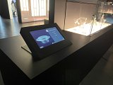 L'image montre un espace d'exposition dans un musée ou une galerie. Au premier plan, il y a un écran tactile sur une table noire qui semble fournir des informations sur les objets exposés. Derrière, on peut voir une vitrine avec des objets en verre, probablement des œuvres d'art ou des pièces de collection. L'éclairage est conçu pour mettre en valeur ces objets transparents et reflétants, créant une ambiance moderne et sophistiquée.