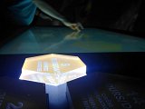 L'image montre une table interactive éclairée, sur laquelle une main semble toucher une surface tactile. Au premier plan, un objet hexagonal lumineux affiche le mot "JOURS" suivi du chiffre "5". La scène est plongée dans une ambiance sombre, ce qui met en valeur les éléments lumineux. Des inscriptions ou des chiffres supplémentaires sont visibles autour de l'objet central, suggérant un environnement éducatif ou muséal.