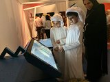 L'image montre deux enfants portant des vêtements traditionnels, probablement des kandouras, en train d'interagir avec un écran tactile. Ils semblent engagés et souriants. À l'arrière-plan, on peut voir une foule de personnes, dont une femme vêtue d'une abaya qui les regarde. L'ambiance semble être celle d'un événement culturel ou éducatif, avec un décor coloré.