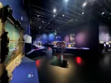 L'image montre un intérieur d'exposition dans un musée ou un centre culturel. Les murs sont peints en bleu foncé, créant une atmosphère moderne et immersive. On aperçoit plusieurs objets exposés sur des vitrines et des tables, dont un cadre doré avec une œuvre d'art. Des éléments lumineux portent l'attention sur certaines pièces, et des informations en texte peuvent être visibles sur les murs. L'éclairage est subtil et met en valeur les objets tout en contribuant à l'ambiance générale de l'espace.
