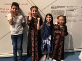 L'image montre quatre enfants qui posent ensemble. Deux filles portent des robes traditionnelles ornées de motifs floraux, tandis qu'un garçon est habillé en t-shirt et pantalon. La fillette au centre porte une robe bleu foncé avec un motif. Tous les enfants affichent un sourire et montrent leur poignet, où ils portent des bracelets blancs. L'arrière-plan contient des inscriptions en plusieurs langues, suggérant un contexte culturel ou artistique.