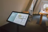 L'image montre un intérieur de musée ou d'exposition. À l'avant-plan, il y a un écran tactile qui présente des informations sur une chaise, probablement une œuvre de design. À l'arrière-plan, on aperçoit une pièce avec une fenêtre ronde et un design intérieur minimaliste, où se trouvent une autre chaise et une table en bois. L'environnement semble moderne et épuré.