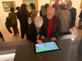 L'image montre une scène à l'intérieur d'un musée. Deux personnes, un homme et une femme, sont en train d'interagir avec un écran tactile qui affiche une œuvre d'art. Ils semblent intéressés par ce qu'ils voient. En arrière-plan, d'autres visiteurs admirent les tableaux accrochés aux murs. L'ambiance est vivante et culturelle, typique d'une exposition artistique.