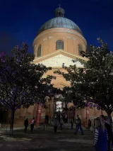 L'image montre un bâtiment impressionnant avec un dôme au sommet, dans une atmosphère nocturne. La façade est de couleur orange et semble bien éclairée, tandis que des arbres sont visibles au premier plan. Des gens se déplacent autour du bâtiment, créant une ambiance animée. Des lumières colorées ajoutent une touche festive à la scène, et le ciel est teinté de nuances sombres, renforçant le contraste avec la structure architecturale.