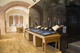 L'image montre un intérieur de musée avec un sol en bois et des murs en pierre. On y voit plusieurs vitrines en verre, aménagées pour exposer des objets ou des documents. Des tables en bois, soigneusement disposées, accueillent des présentations thématiques. La lumière naturelle illumine l'espace, créant une ambiance chaleureuse et accueillante. L'architecture est à la fois moderne et traditionnelle, avec des arches décoratives visibles en arrière-plan.