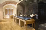 L'image montre un intérieur de musée avec un sol en bois et des murs en pierre. On y voit plusieurs vitrines en verre, aménagées pour exposer des objets ou des documents. Des tables en bois, soigneusement disposées, accueillent des présentations thématiques. La lumière naturelle illumine l'espace, créant une ambiance chaleureuse et accueillante. L'architecture est à la fois moderne et traditionnelle, avec des arches décoratives visibles en arrière-plan.