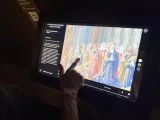 L'image montre une personne interactant avec un écran tactile qui affiche une œuvre d'art. Sur l'écran, on peut voir une scène colorée représentant des figures humaines, probablement des personnages historiques ou bibliques, dans un style médiéval. Les détails des vêtements et des visages suggèrent une période ancienne, et l'arrière-plan est orné de motifs architecturaux ou décoratifs. La personne semble être engagée dans l'exploration ou l'apprentissage de l'œuvre présentée.