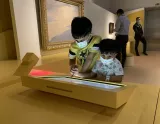 L'image montre deux enfants dans un musée, interagissant avec une tablette ou un écran tactile situé sur une table jaune. Les enfants portent des masques et semblent concentrés sur ce qu'ils manipulent. En arrière-plan, on peut apercevoir d'autres visiteurs et des œuvres d'art accrochées aux murs. L'environnement est moderne et lumineux.